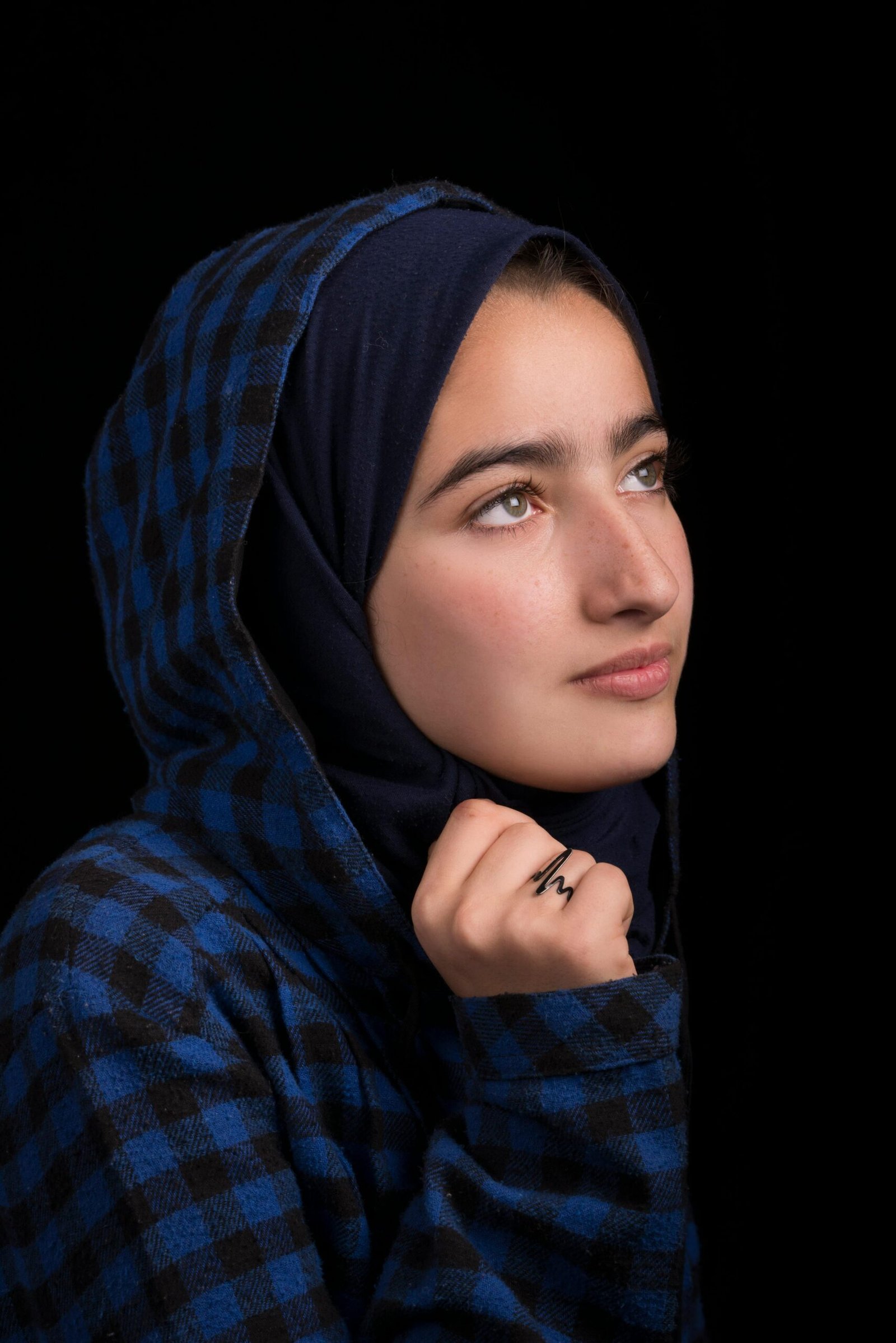 A close-up portrait of a woman wearing a blue plaid hijab, looking thoughtful against a dark background.